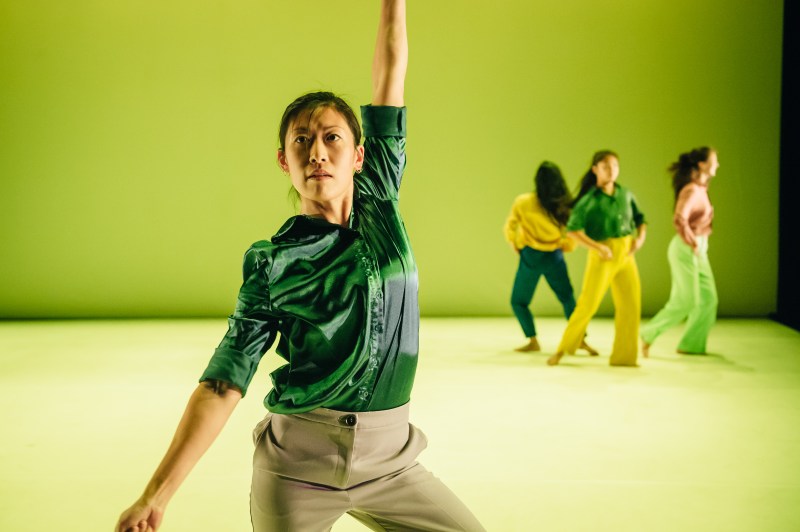 In the foreground is a female dancer in a shiny turquoise shirt and light coloured trousers eyes looking up with her arms pulled apart in opposite directions. Behind her are three female dancers with their hands on their waists in the motion of turning. They are in colour coordinated shirts and trousers with long hair loose.